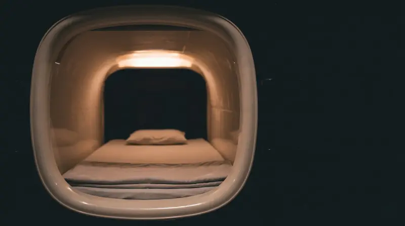 A softly lit sleeping pod with a thin mattress and pillow set against a dark surround.